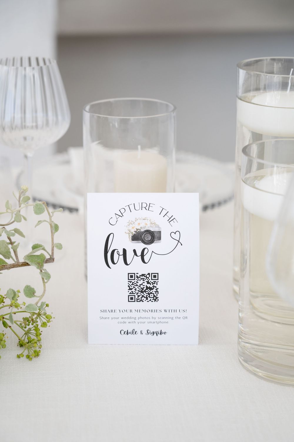 Wedding reception card with camera logo and QR code for sharing memories sits between glasses on white table.