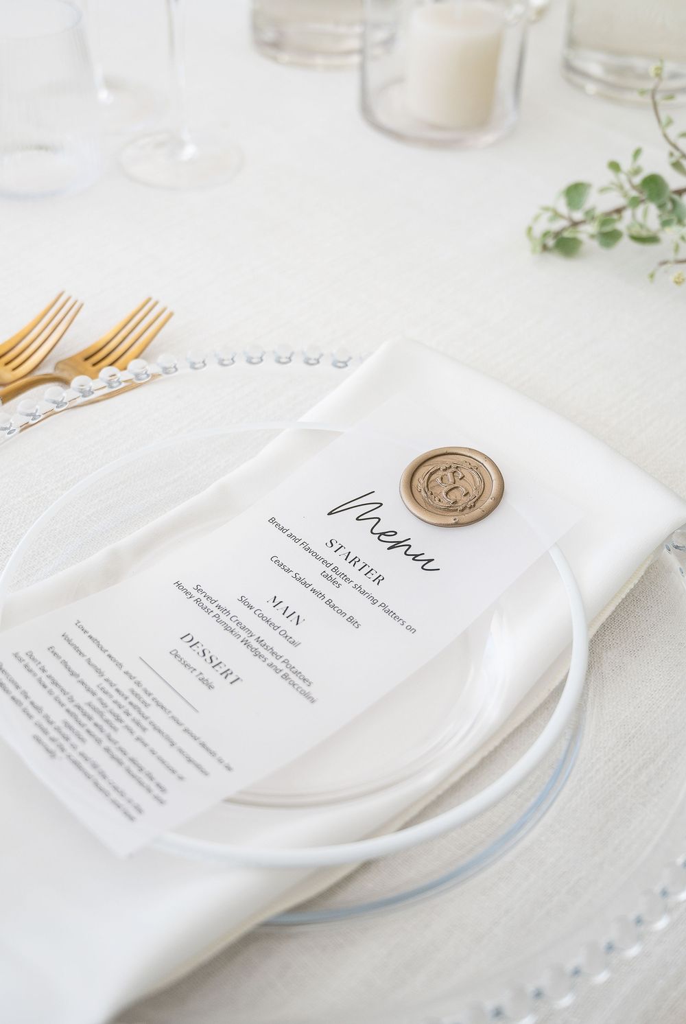 Elegant white place setting with gold cutlery and a wax-sealed menu card on a crisp white tablecloth setting.