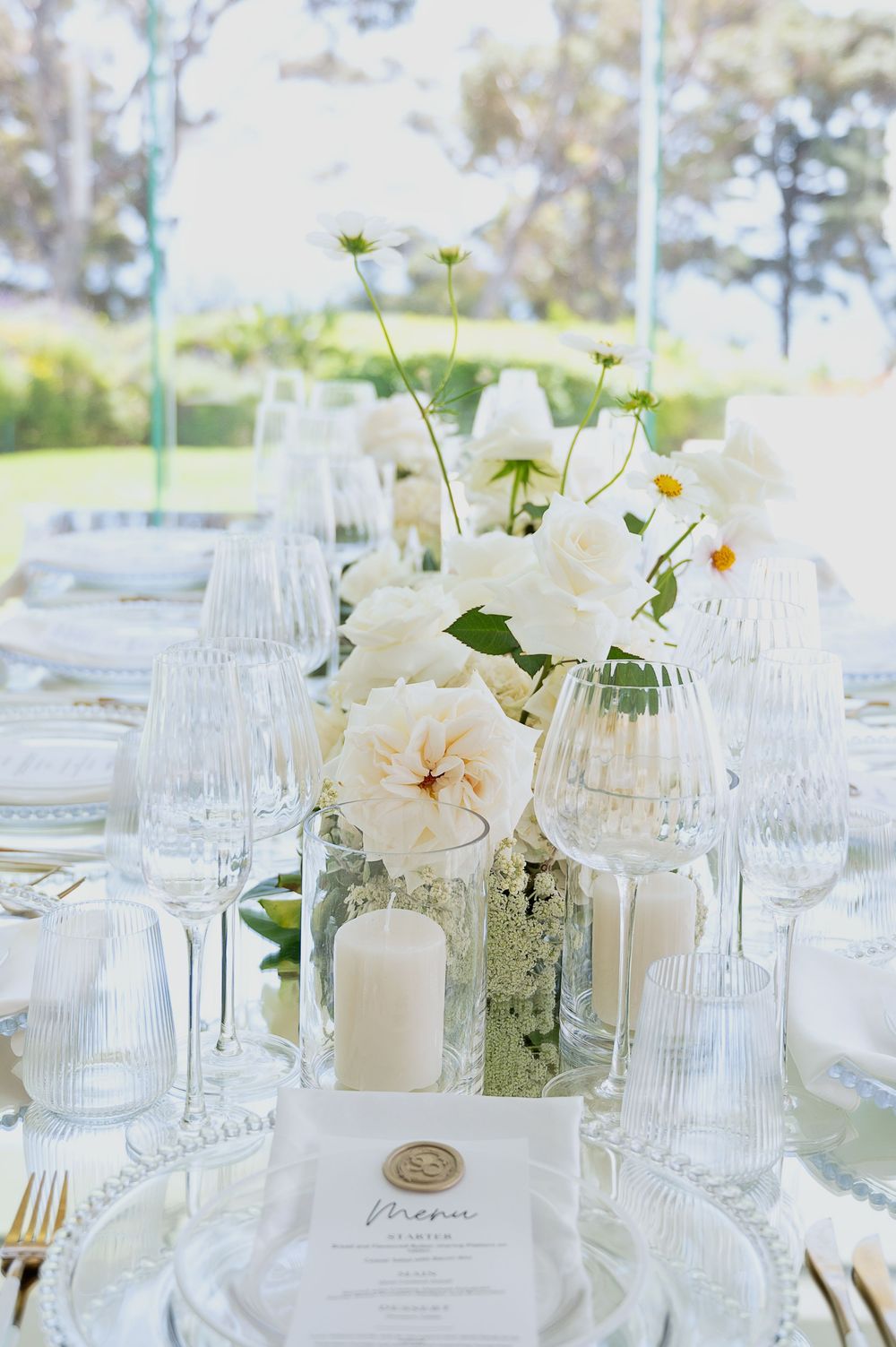Elegant white wedding reception table settings with glass stemware, floral centerpieces and white candles in ethereal light.