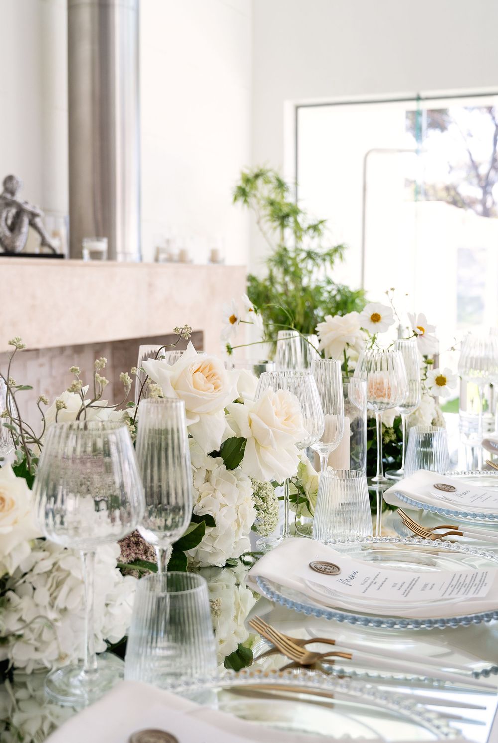 Elegant white wedding reception tablescape with glass vases, delicate flowers and silver accents in a bright sunlit room.