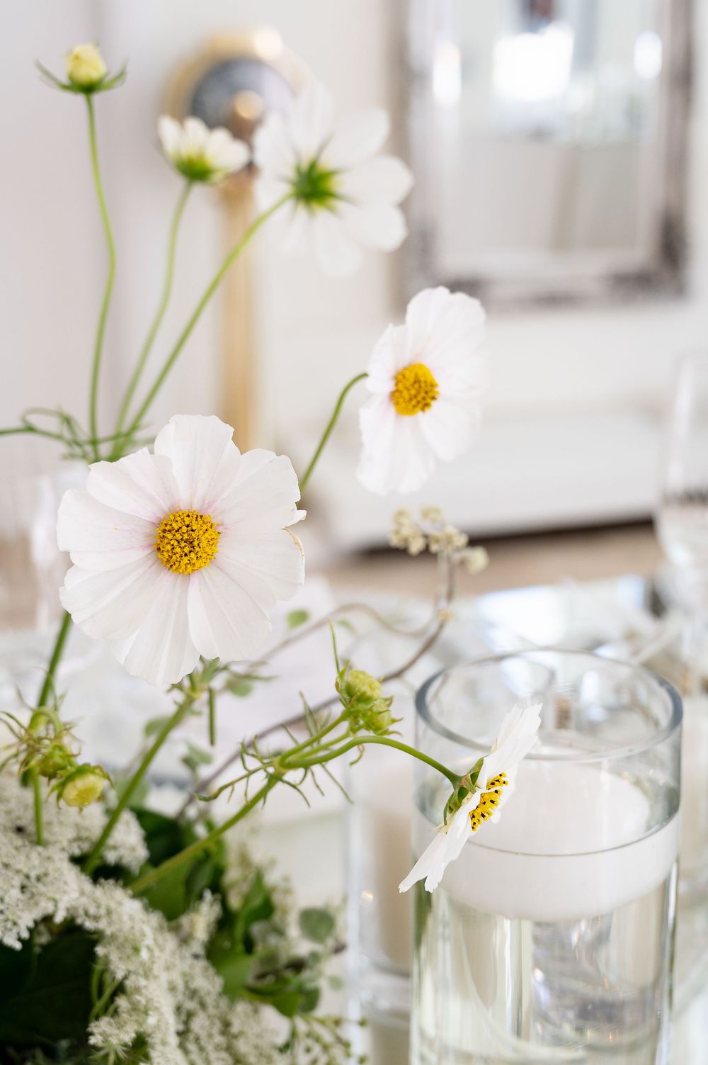 White cosmos flowers with yellow centers arranged in a glass vase alongside white candles on an elegant tablescape.