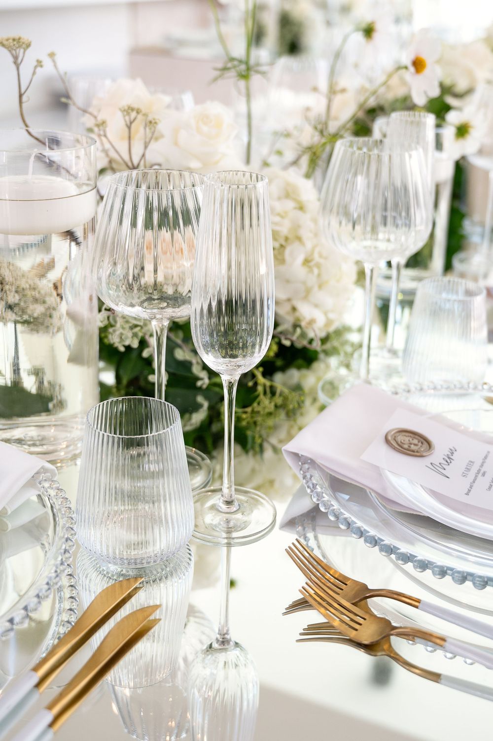 Elegant table setting with white flowers, crystal glassware, golden cutlery and delicate china plates in a luxurious style.