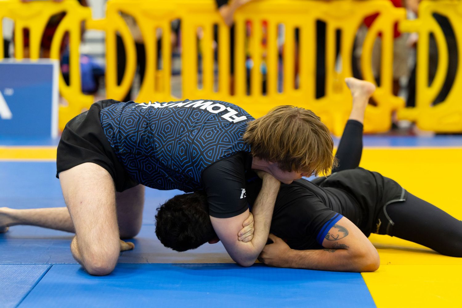Competitors engaged in ground fighting techniques on a yellow competition mat.