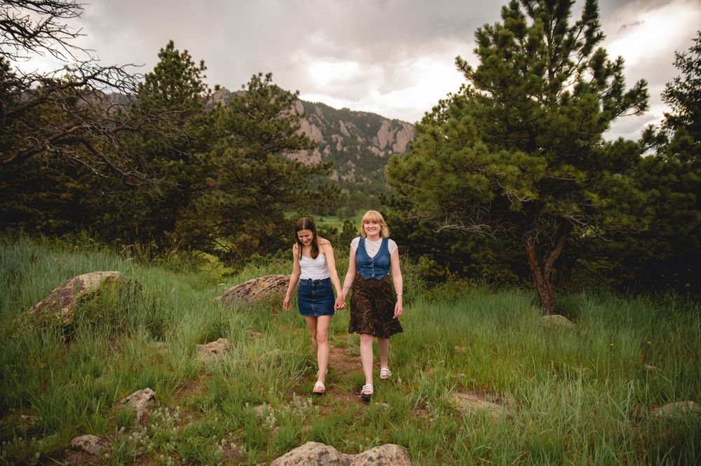 People hiking together along scenic mountain trail through tall green grass and pine trees at sunset.