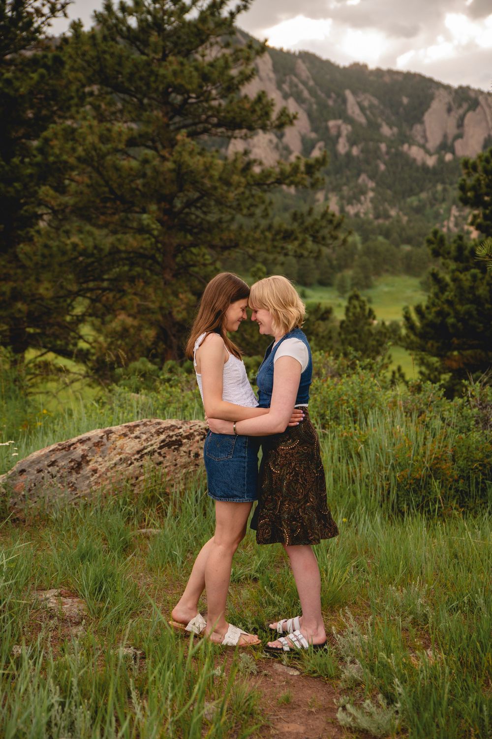 A couple sits on a rocky hillside enjoying a romantic moment against mountain scenery with green grass and trees.