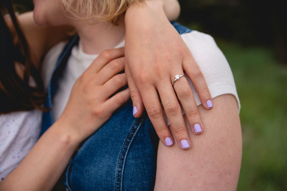 Close-up view of hand with pale purple nail polish resting on denim-clad knee, showing delicate engagement ring.