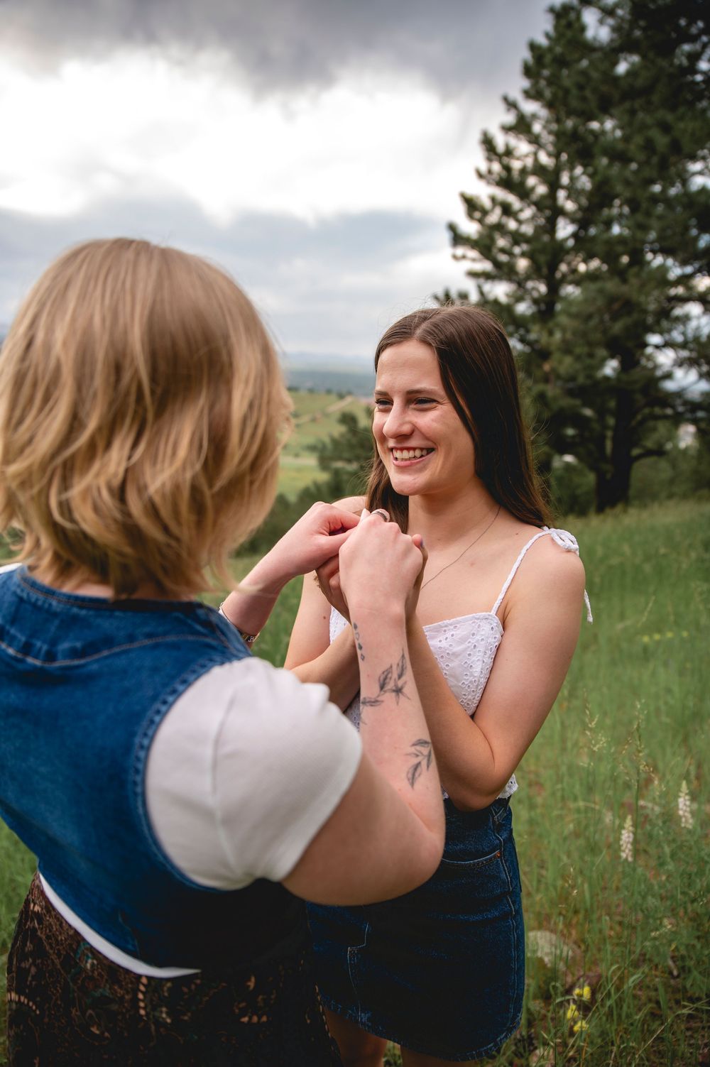 Two friends sharing laughter and joy while sitting in a grassy meadow with mountain views and evergreen trees.