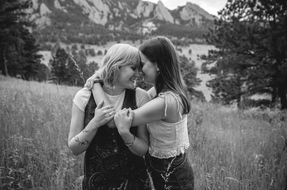 Two friends share an intimate moment together in a grassy meadow with majestic mountain peaks in the background.