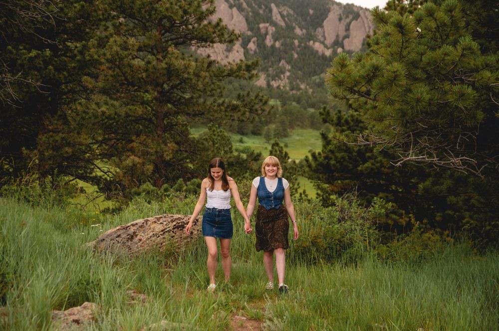 Two people walk together on a mountain trail surrounded by green grass and trees with rocky cliffs in the background.