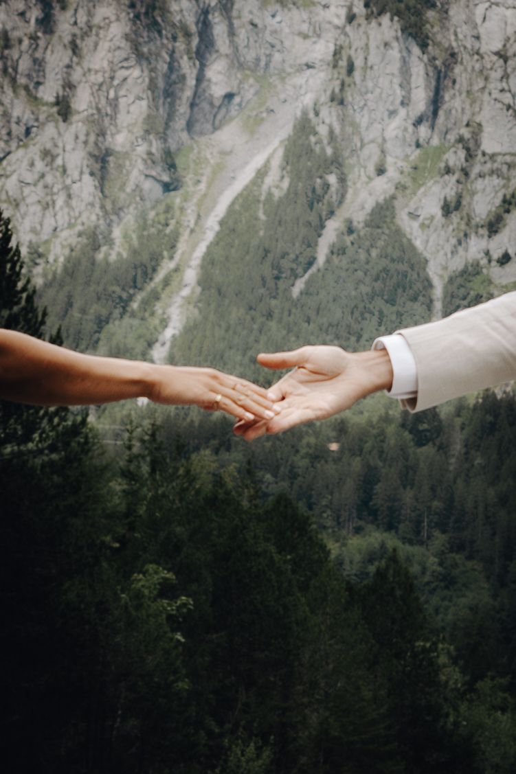 Two hands reaching towards each other with a rugged mountain landscape and evergreen forest in the background.