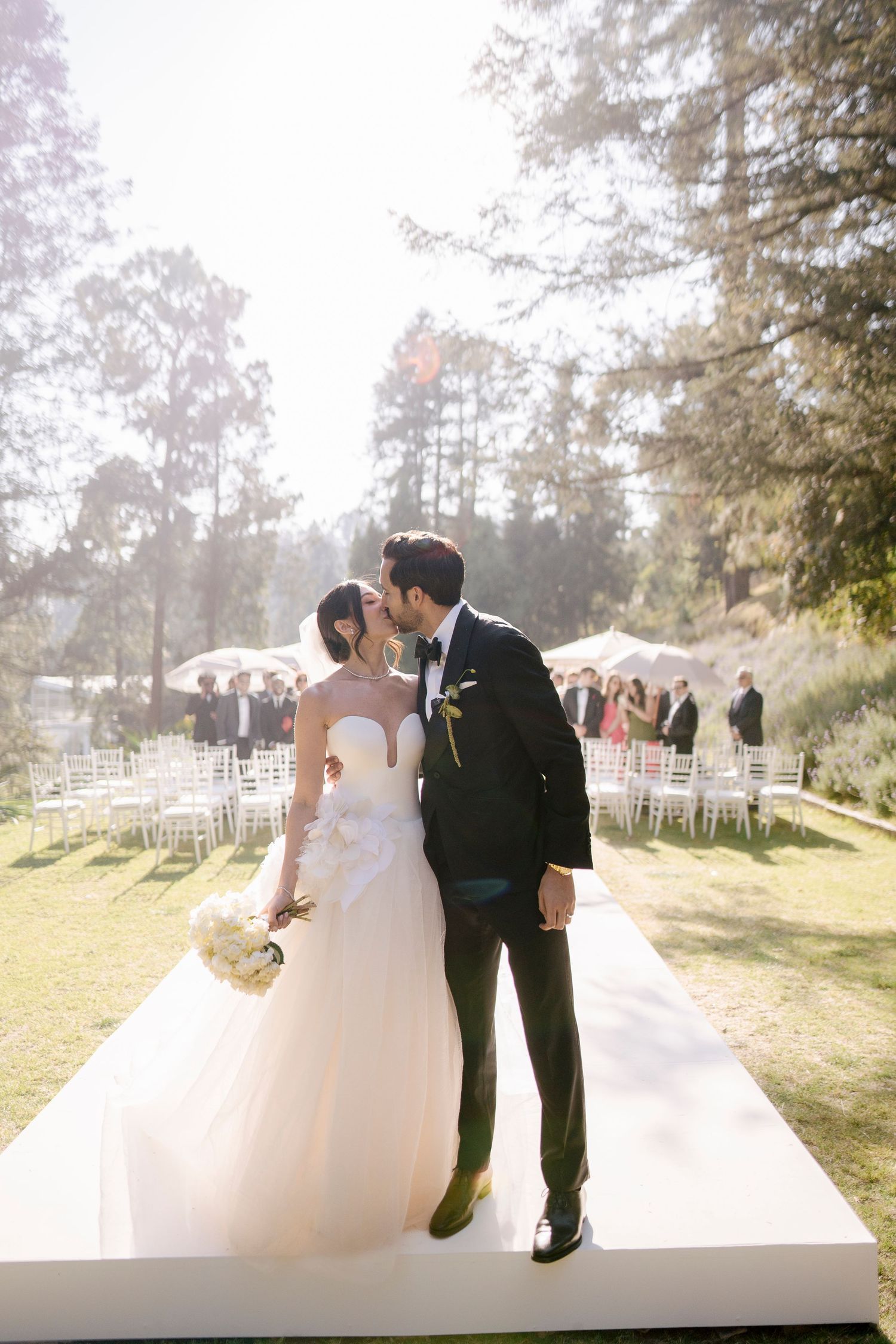 Outdoor wedding gathering on a lawn with mountain backdrop and white chairs arranged in foreground at sunset.