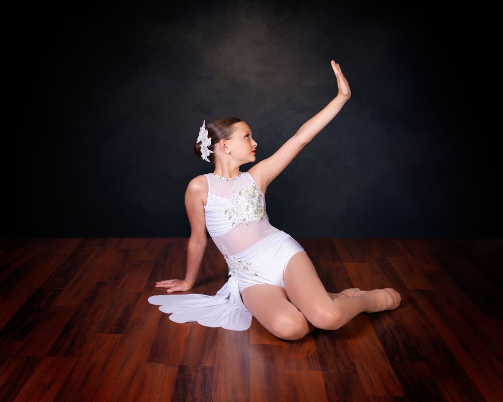 Dancer in white costume strikes graceful pose on dark wooden floor with dramatic black background.