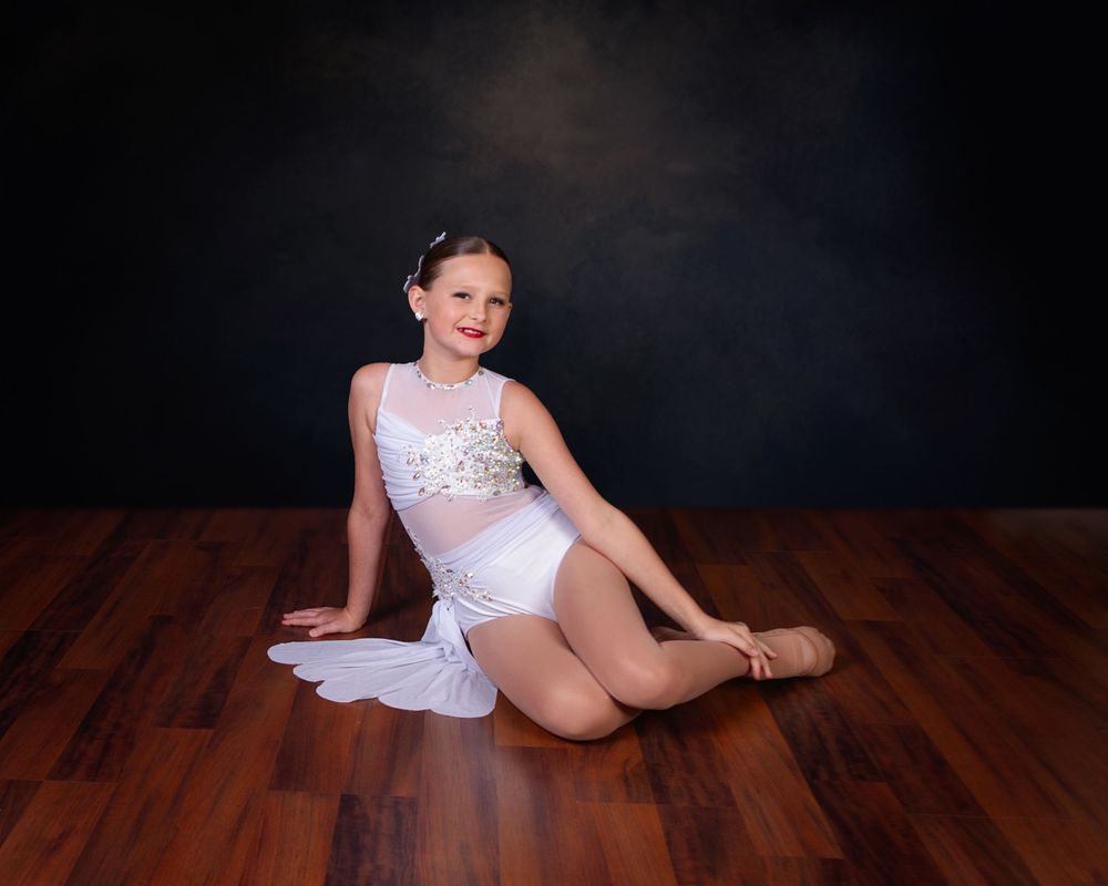 Young dancer in white leotard and sheer overlay poses gracefully on wooden floor against dark studio backdrop.