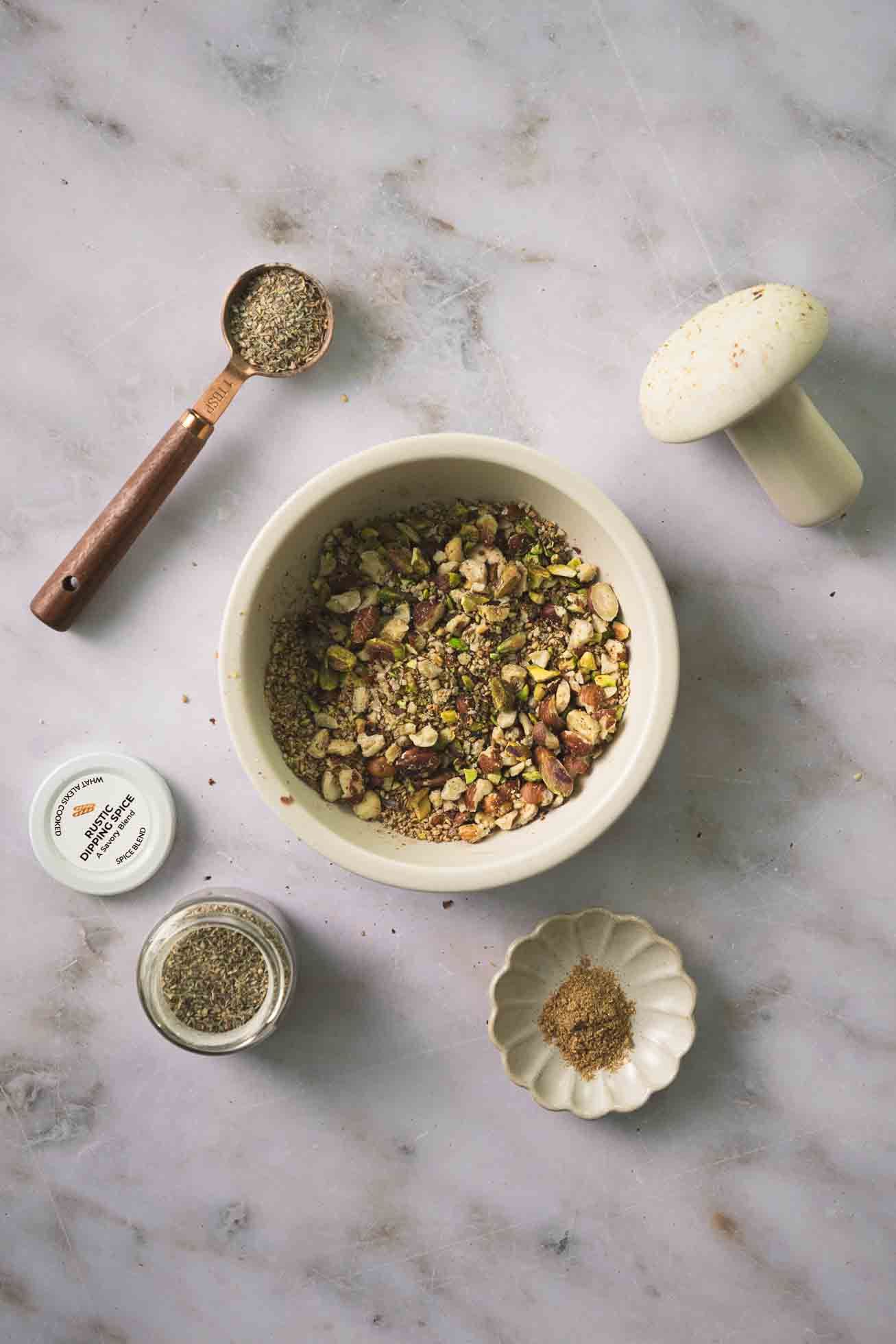 Overhead view of dried herbs and spices in small bowls and measuring spoons on marble surface.