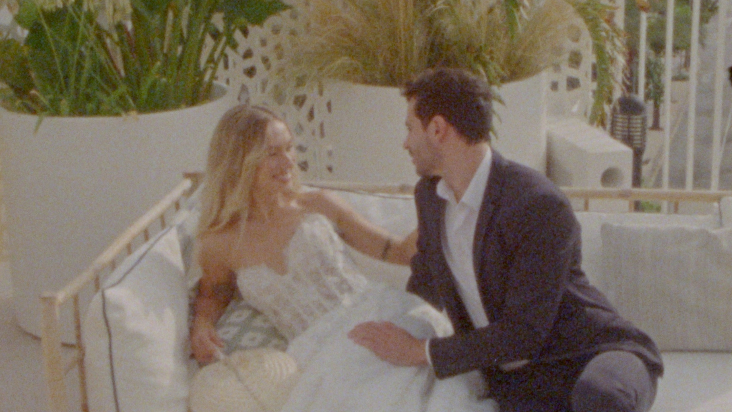 A couple in formal wedding attire sits together on an outdoor white couch having an intimate conversation.