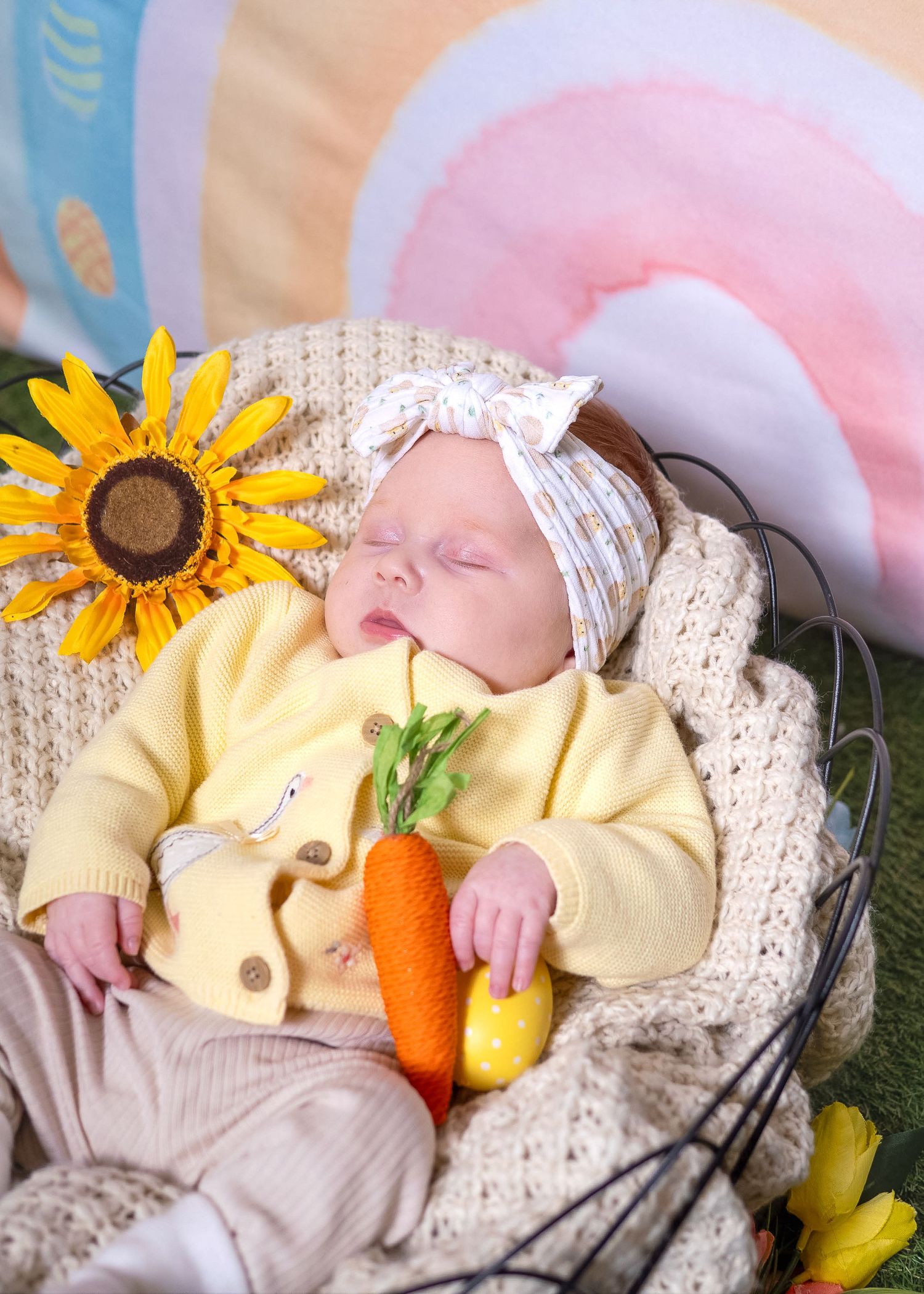Easter Mini Session - A sleeping baby in yellow clothing lies in a basket with decorative carrots and sunflowers.