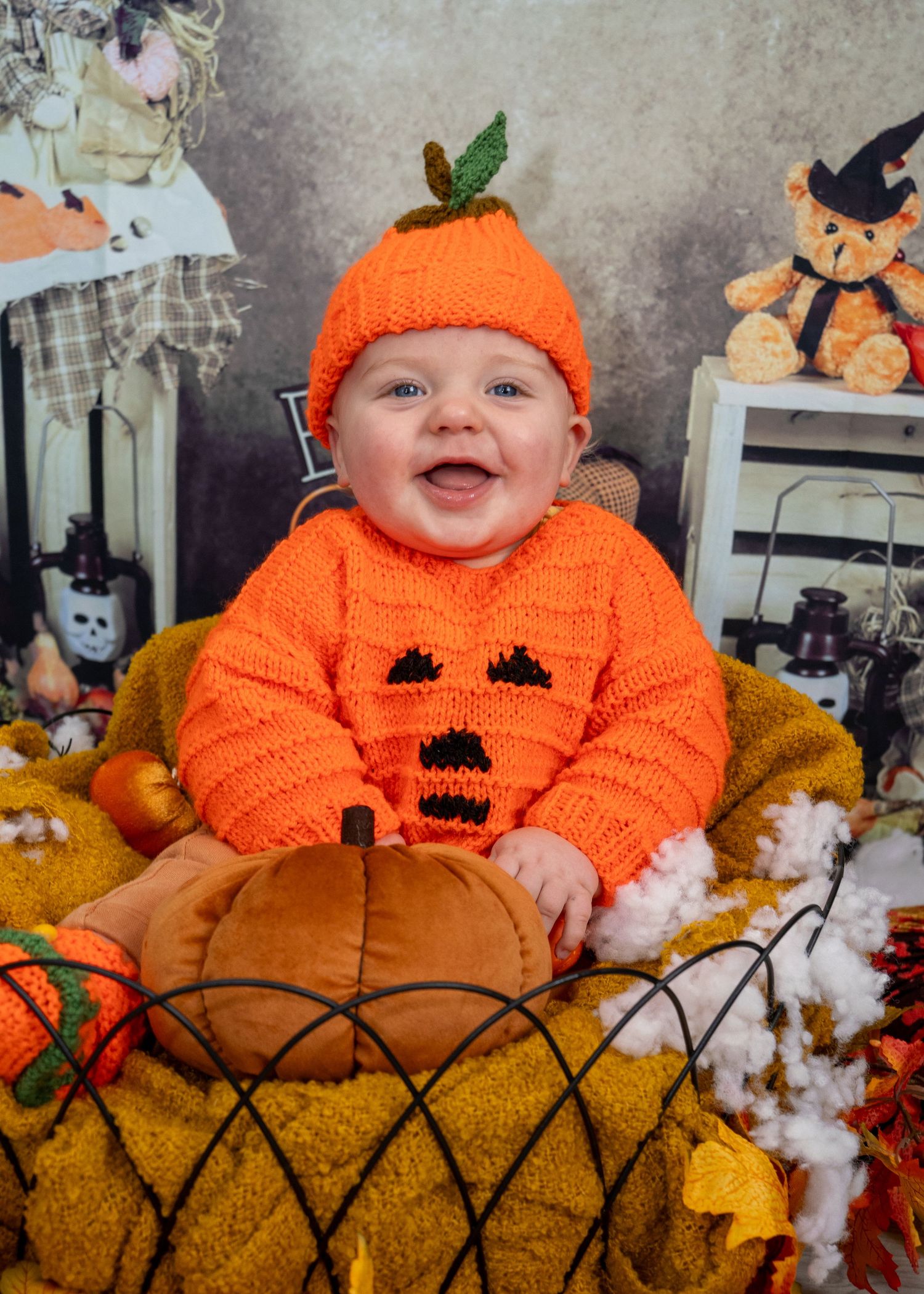A baby in an orange pumpkin costume poses on a yellow couch with Halloween decor.