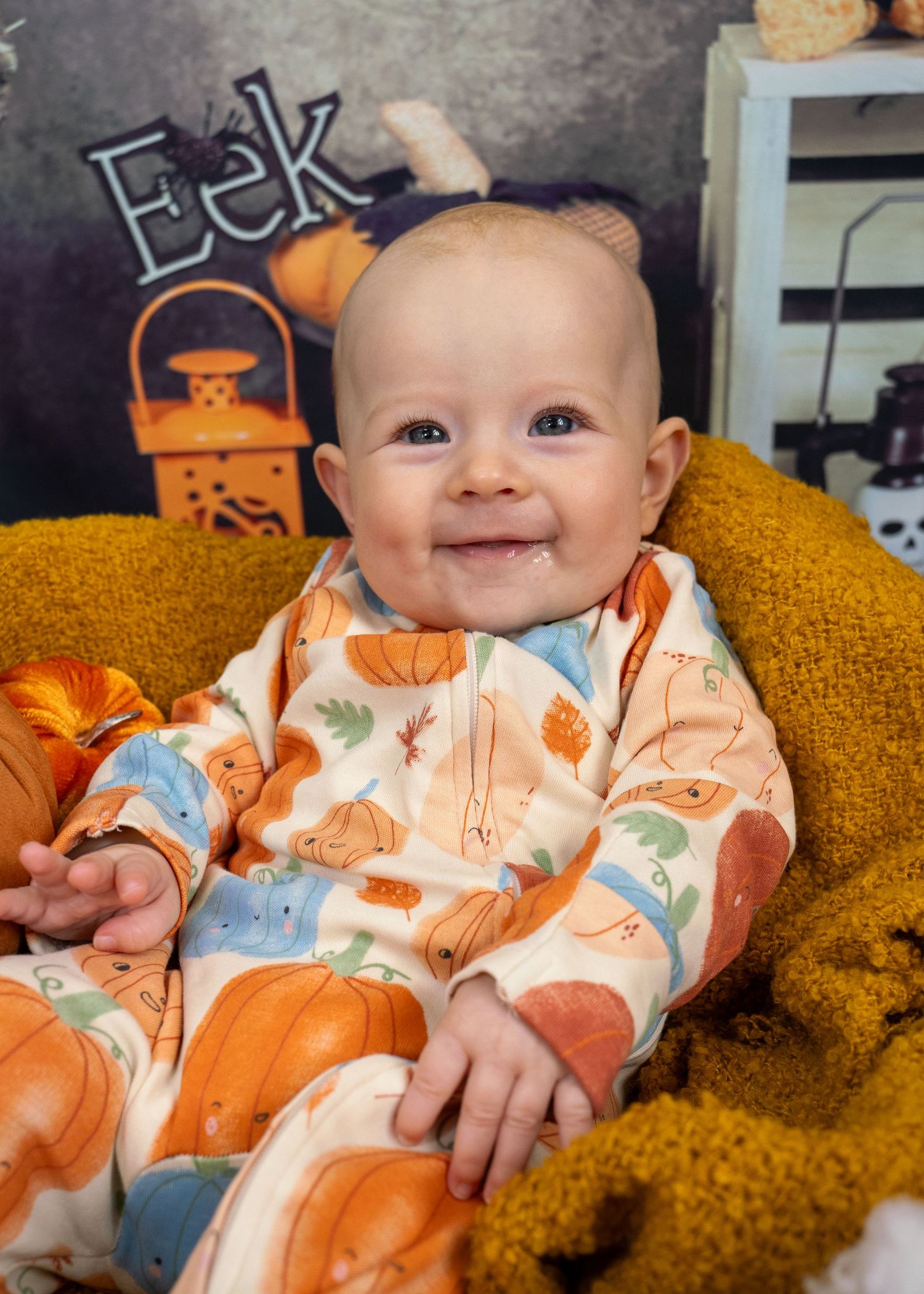 A baby in pumpkin-print pajamas smiles while sitting on a mustard yellow couch.