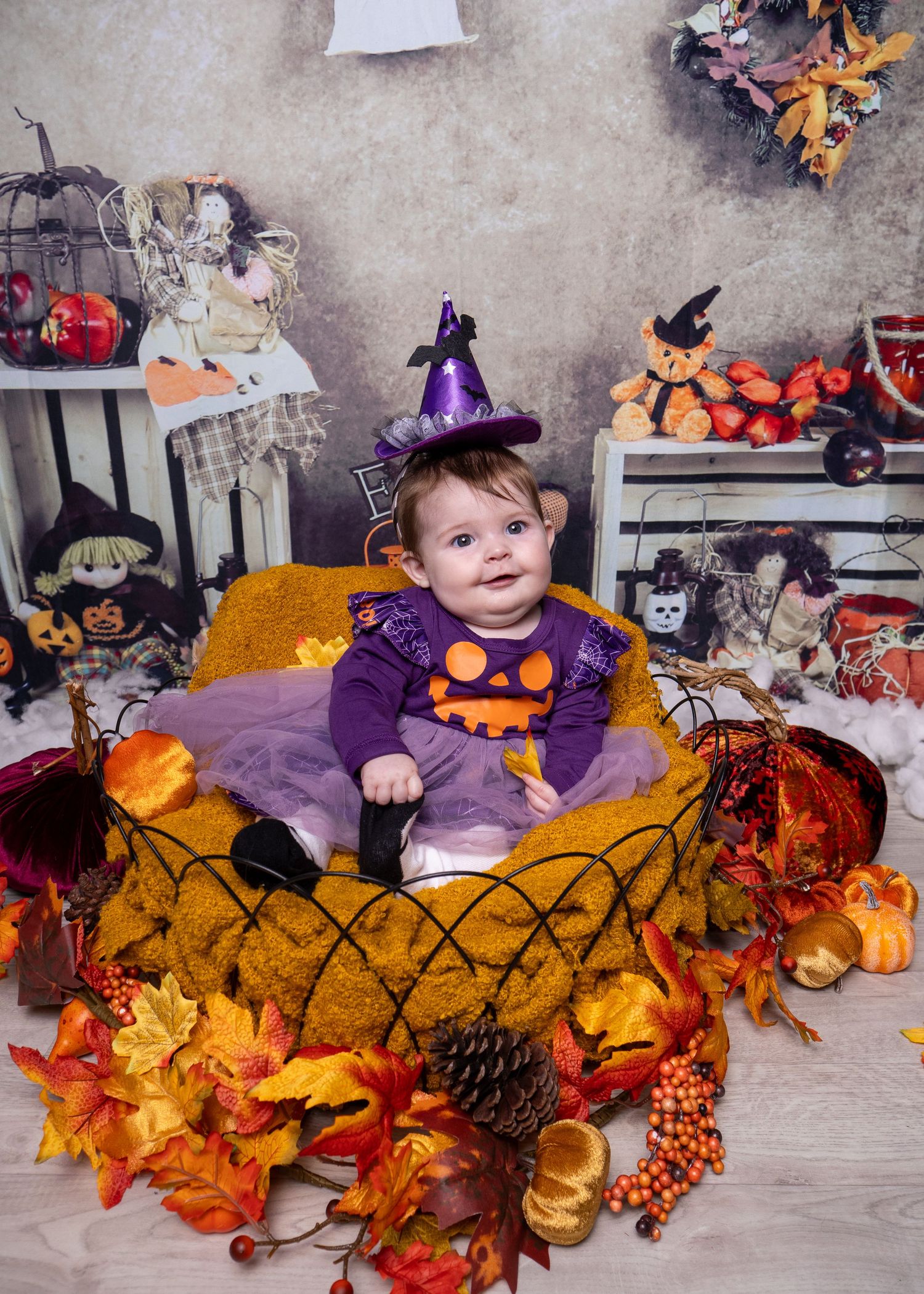 Autumn themed baby photo setup with pumpkins, leaves, and Halloween decorations in purple and orange tones.