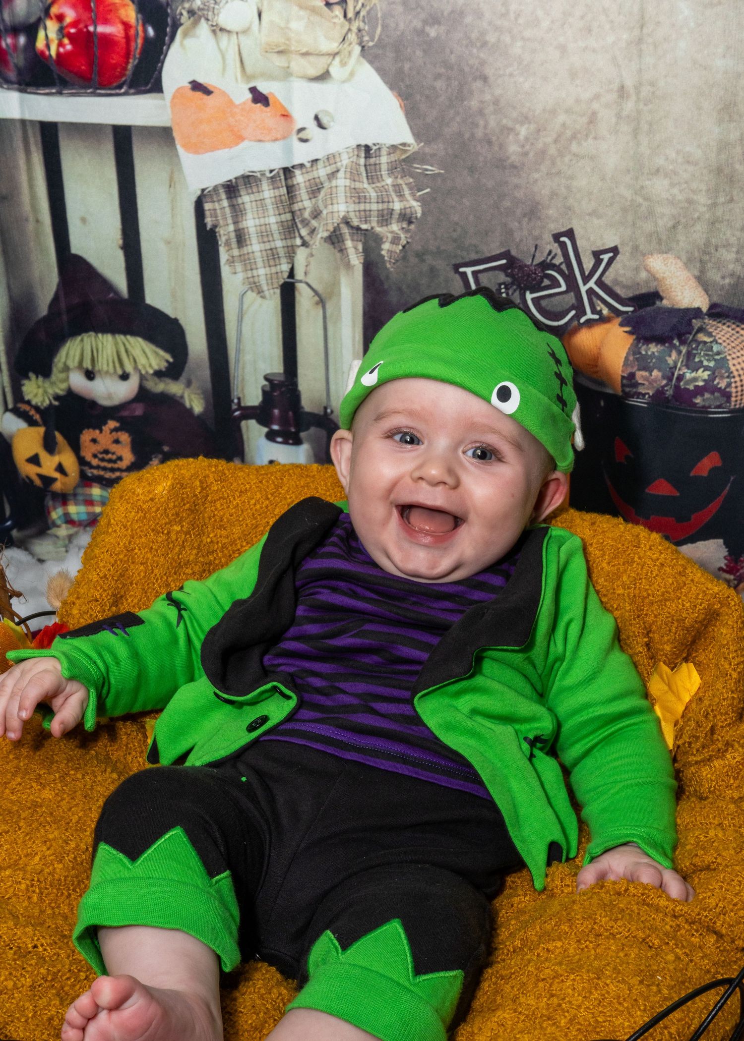A baby wearing a green costume smiles while sitting on a yellow couch with Halloween decorations.