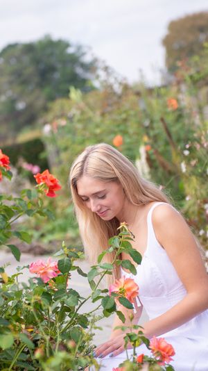 Senior in white dress admires blooming red roses in garden setting.