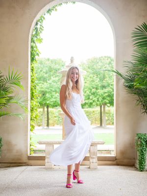 High School senior in white dress dances under arched walkway with pink shoes.