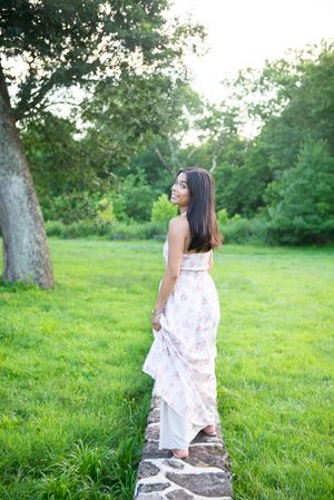 Woman in flowing floral dress stands on stone wall in lush garden setting at sunset.