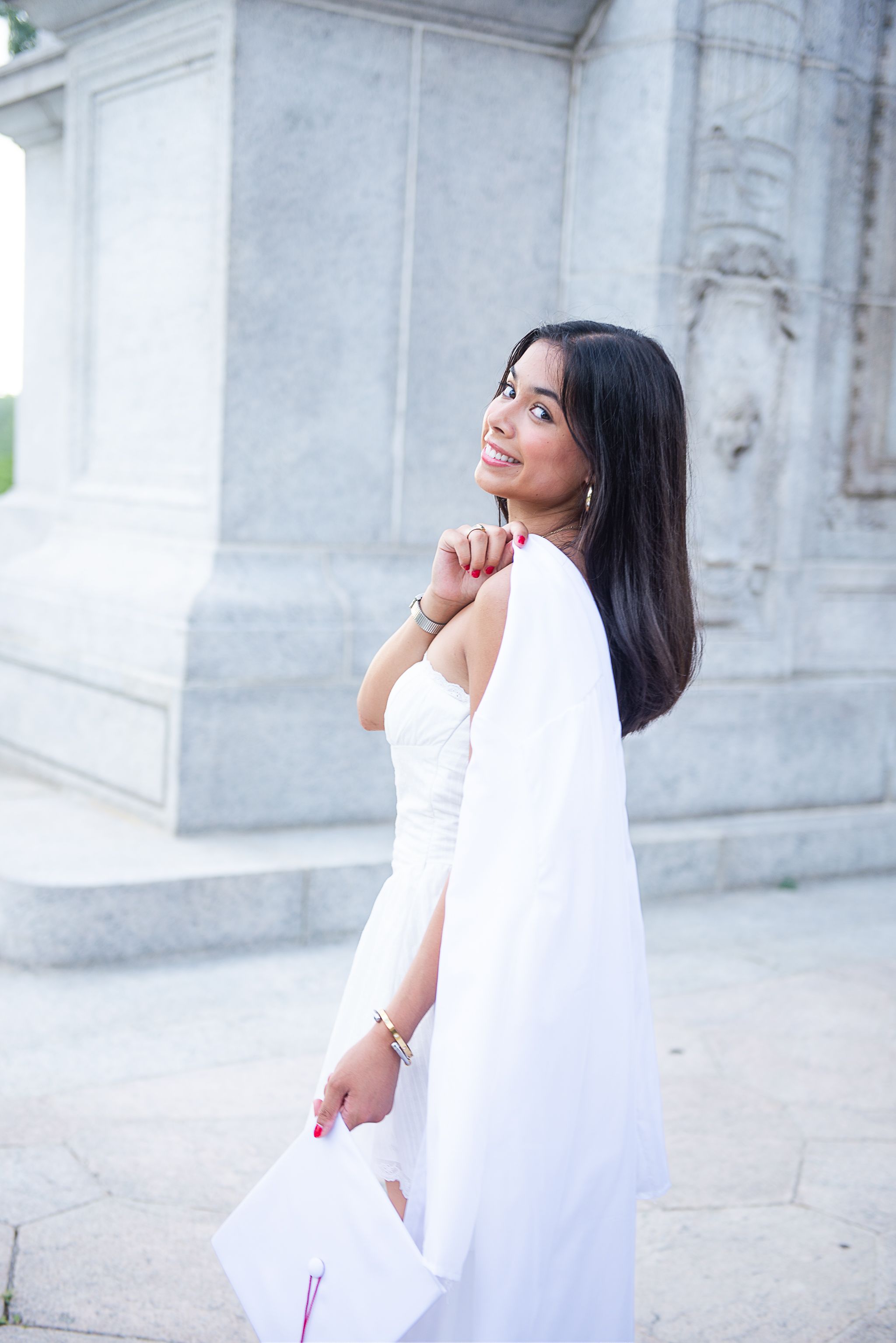 Graduate in white dress holds diploma while posing against marble columns.