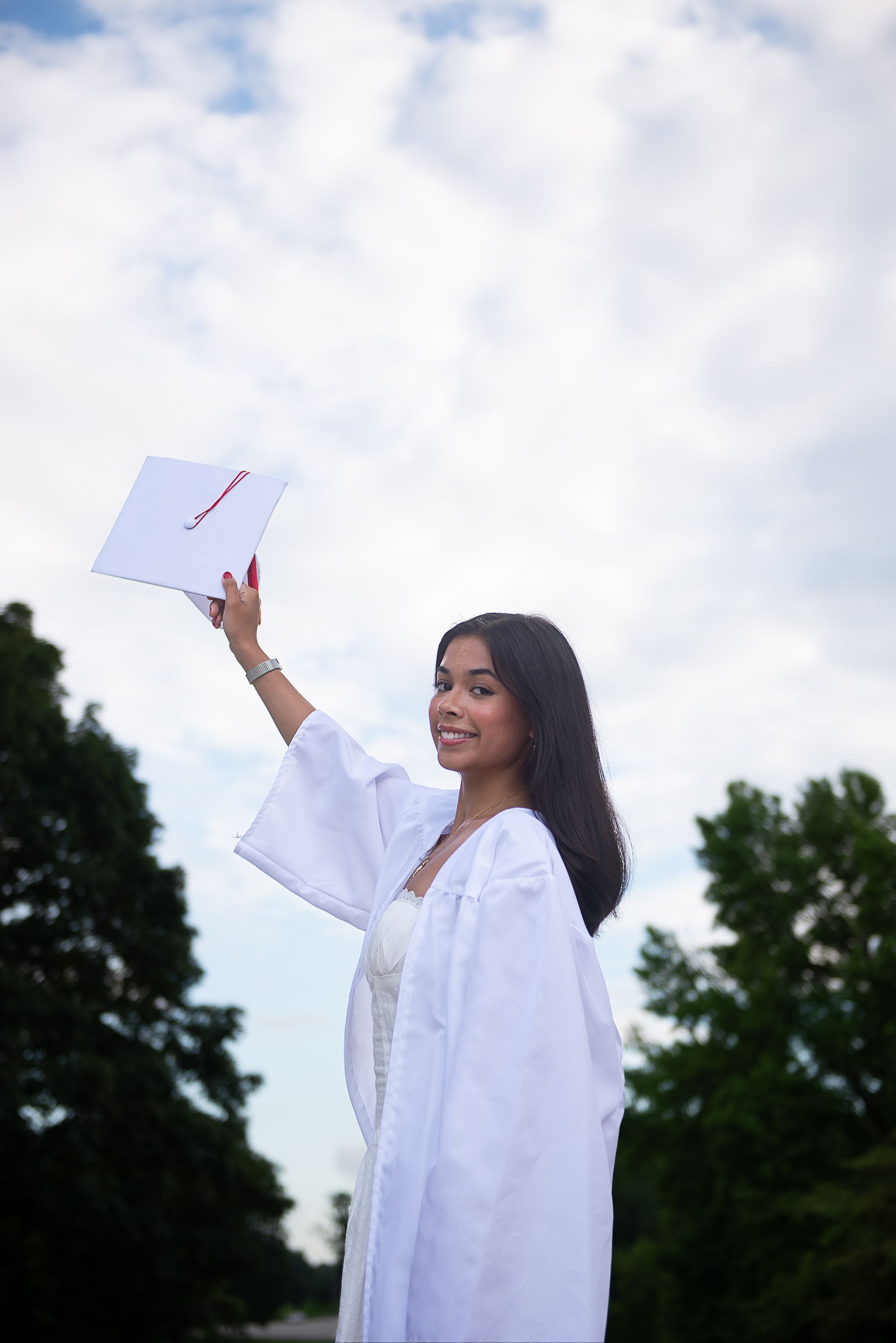 Graduate in white gown holds graduation cap up against cloudy sky.