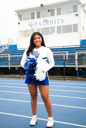 Cheerleader in blue and white uniform holding pom poms on track field with Patriots stadium backdrop.