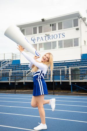Cheerleader in blue and white uniform performs on blue track field with stadium in background.