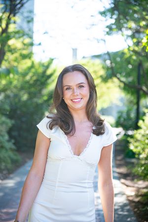 Woman in white blouse smiles outdoors with natural greenery in background.