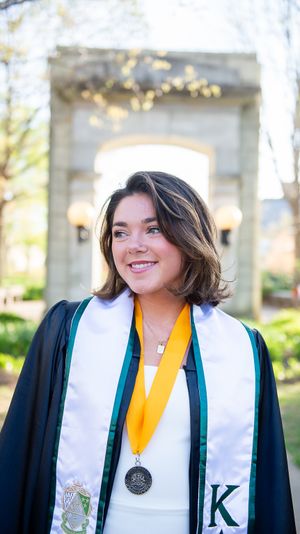 Graduate in academic regalia with gold honor cord stands in front of stone archway.