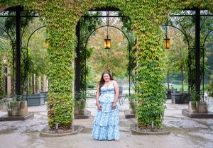Person in blue floral dress poses under ivy-covered garden archway.