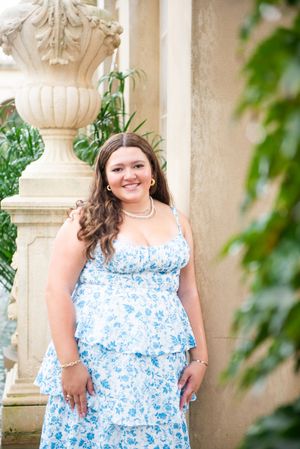 Portrait in blue floral dress standing by ornate stone column in garden setting.