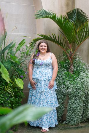 Person in blue floral maxi dress standing among tropical plants and palm fronds.
