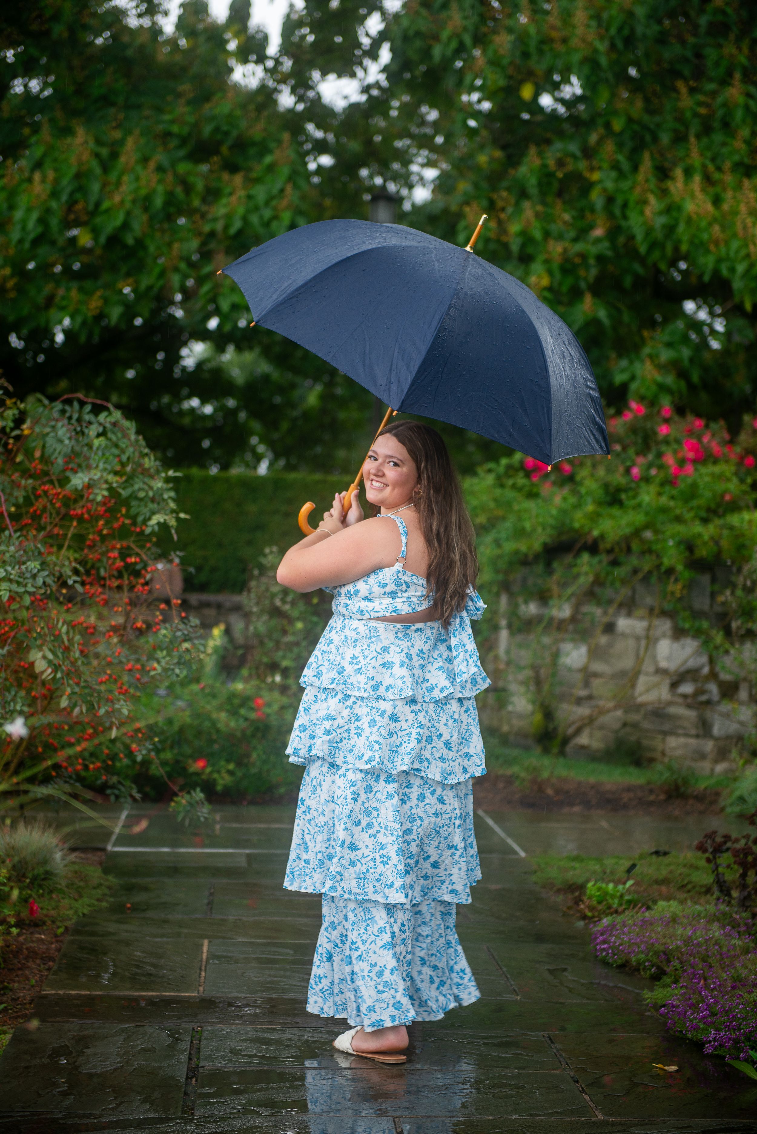 Person in floral dress twirls with umbrella in garden during rainy photoshoot.