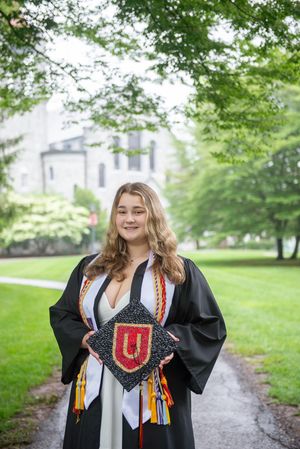 Graduate in black cap and gown holding decorated mortarboard with U emblem on campus grounds.
