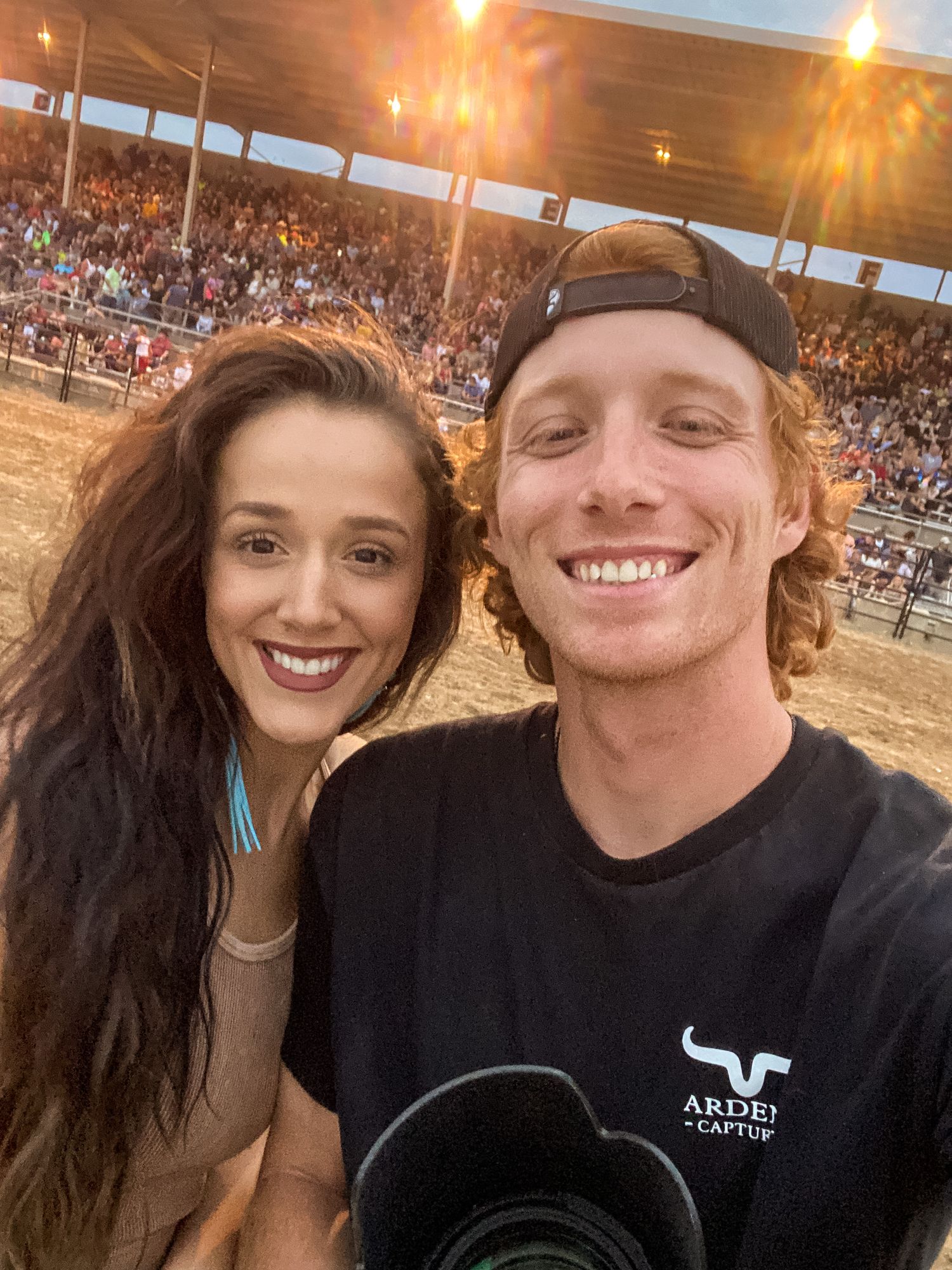 A couple takes a selfie at a crowded rodeo arena during sunset with stadium lights glowing in the background.