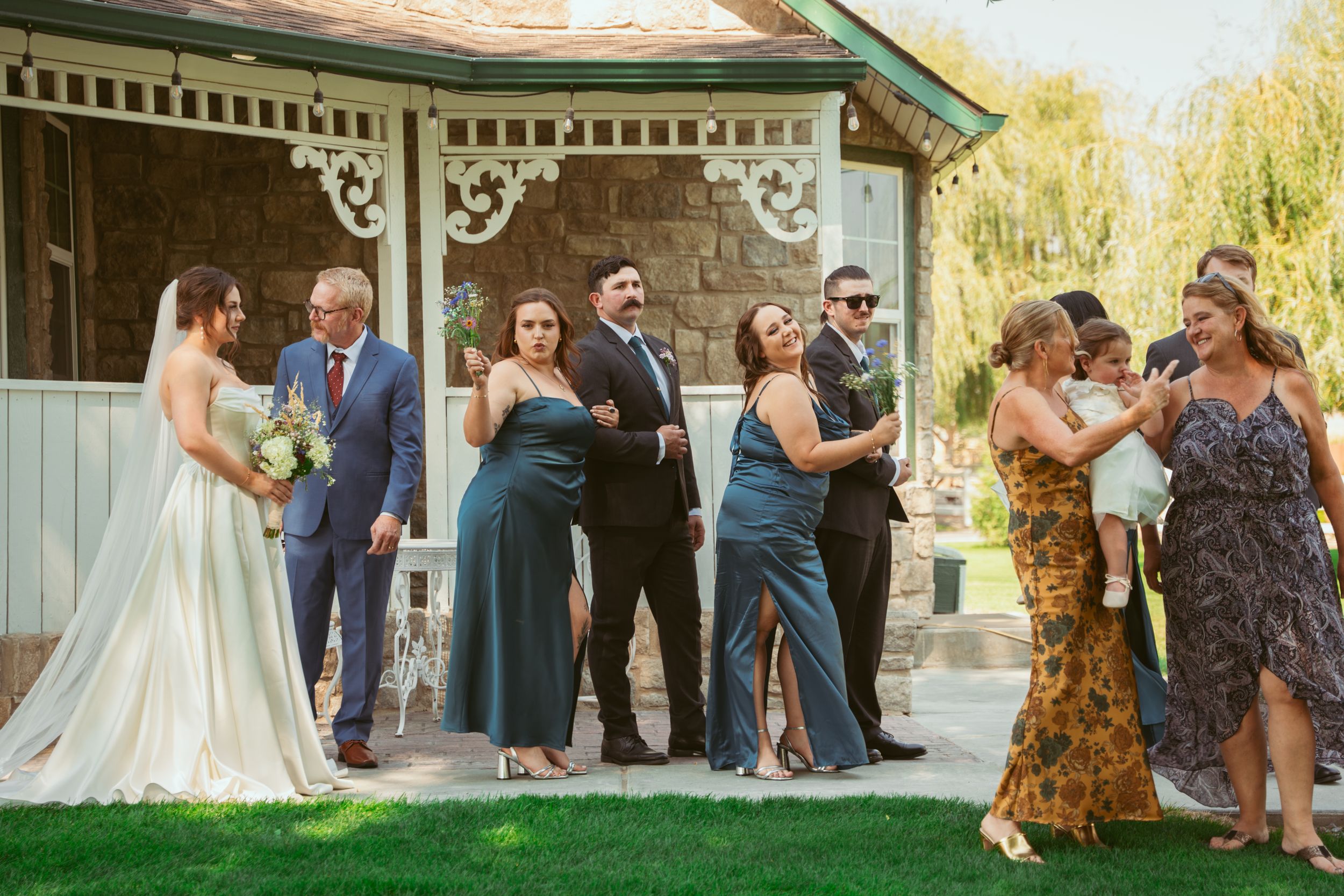 Wedding party gathers under a willow tree for an outdoor ceremony in warm sunlight.