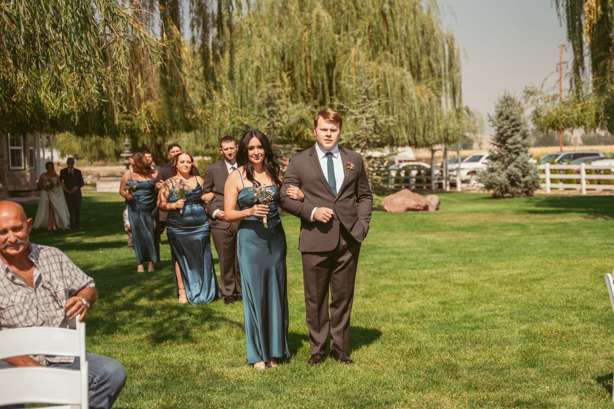 Wedding party members walk down a grassy aisle lined with weeping willow trees on a sunny outdoor ceremony.