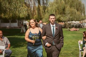 Members of a bridal party process down an outdoor ceremony aisle with weeping willows in the background.