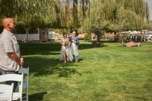 People gather on a sunny lawn with white chairs set up for an outdoor event with weeping willow trees in the background.