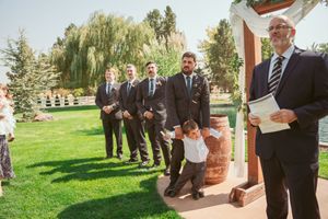 Groomsmen and a young ring bearer line up outside a rustic venue with white chairs and trees in the background.