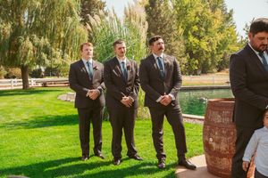 Groomsmen in dark suits stand together on manicured lawn with willow trees and rustic barrel in background.
