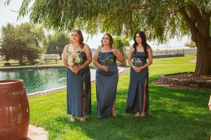Three bridesmaids in matching grey dresses holding floral bouquets pose beside a pond under a willow tree.
