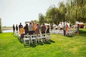 Guests gather on white chairs for an outdoor wedding ceremony under weeping willow trees beside a lake.