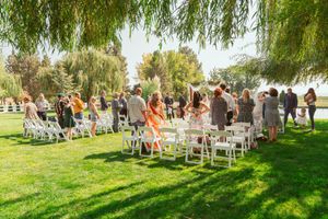 Wedding guests stand among arranged white chairs on a lush green lawn beneath drooping willow branches.