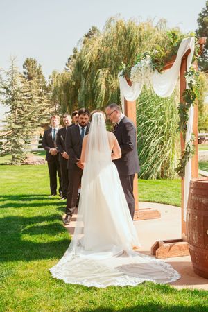 Wedding ceremony takes place outdoors on a sunny day with decorative wooden arch and white flowers.