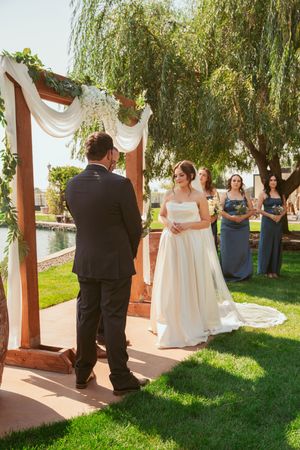 Outdoor wedding ceremony under wooden arch with white fabric draping and weeping willow trees in background.